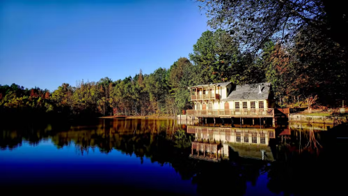A serene lakeside view featuring a wooden house reflecting on calm water, surrounded by trees and a clear blue sky.