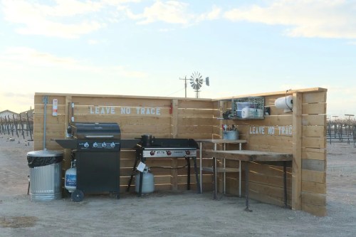 Outdoor cooking area with a grill, table, and trash can, surrounded by wooden fencing and a "Leave No Trace" sign.