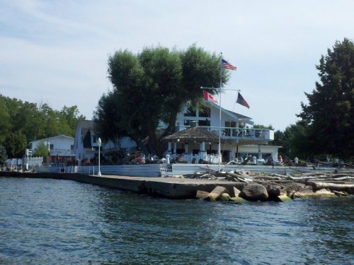 A waterfront view of a large white house with flags, surrounded by trees and a dock on a calm lake.