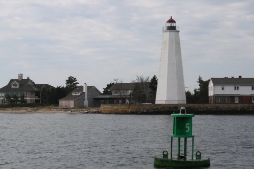 A white lighthouse stands near a shoreline, with green buoy marked "5" in the foreground and houses in the background.