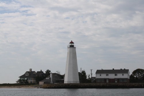 A tall white lighthouse beside two houses, set against a cloudy sky and calm water.