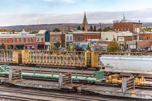 A view of a small town with buildings, a church steeple, and freight train cars in the foreground.