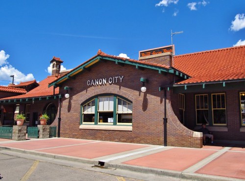 Historic brick train station with a red tile roof, featuring "Canon City" signage and a clear blue sky.