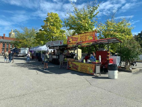 Food trucks and vendor tents at an outdoor market, with trees and a blue sky in the background.