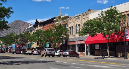A charming downtown street lined with historic buildings, trees, and parked cars, with mountains in the background.