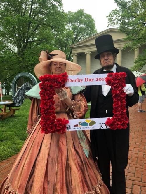 A man and woman in vintage attire pose with a floral frame for Derby Day 2018, set in a park.