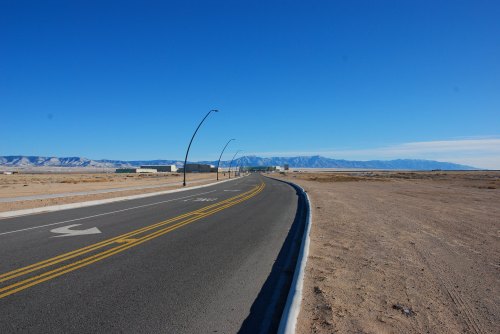 A long, empty road stretches into the distance, flanked by desert terrain and mountains under a clear blue sky.