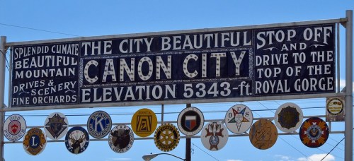 Sign for Canon City, featuring elevation and local organizations' emblems, with a blue sky backdrop.