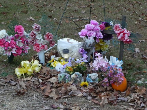 A small memorial with flowers, a cross, and a lantern, surrounded by fallen leaves and a pumpkin.