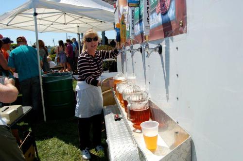 A woman serves beer from a tap at an outdoor event, with people and tents in the background.