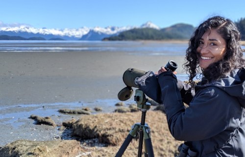 A person with curly hair smiles while using a telescope by a scenic coastal landscape with mountains in the background.
