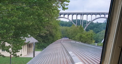 View from a train window showing a bridge in the distance, surrounded by trees and a building in the foreground.