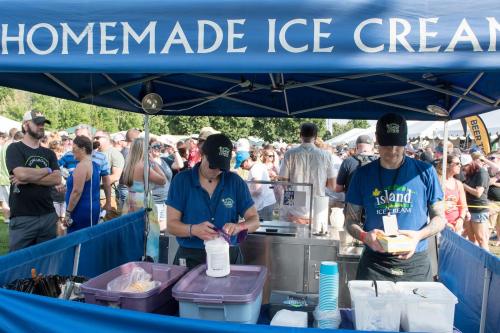 Two vendors serve ice cream at a festival booth, with a crowd of people enjoying the event in the background.