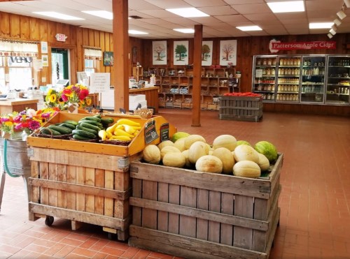 A farm stand interior with wooden crates of melons and squash, colorful flowers, and shelves of cider products.