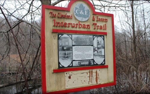 Sign for the Cleveland & Eastern Interurban Trail, featuring historical photos and information, surrounded by trees.