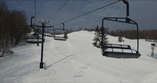A ski lift with empty chairs ascends a snowy slope surrounded by trees under a cloudy sky.