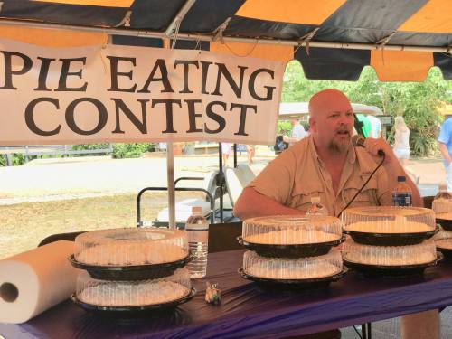 A man sits at a table with pie containers, preparing for a pie eating contest under a tent with a sign.