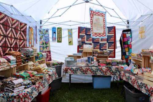 A colorful craft booth featuring quilts, fabric, and handmade items displayed on tables under a white tent.