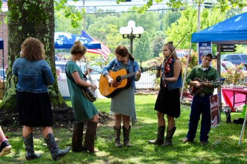 A group of musicians in cowboy boots play instruments outdoors at a festival, surrounded by trees and tents.