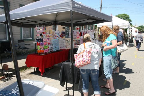 People browsing a vendor's booth at an outdoor market, with colorful crafts displayed under a canopy.