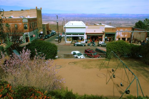 A view of a small town with shops, cars parked, and a playground with swings in the foreground against a mountainous backdrop.