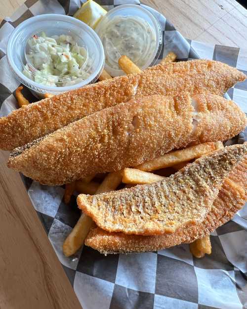 A plate of crispy fried fish fillets with French fries, coleslaw, and dipping sauce on a checkered paper.