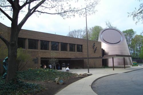 A modern building with a large circular feature, surrounded by trees and greenery, with people near the entrance.