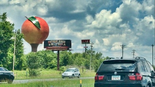 A large peach-shaped water tower stands against a cloudy sky, with cars on a highway and a billboard for antique bikes.
