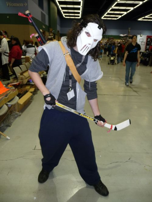 A person in a hockey mask and casual clothing poses with a hockey stick at a convention.