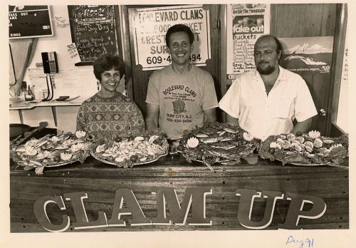 A black-and-white photo of three people behind a seafood counter with trays of clams and crabs, smiling at the camera.