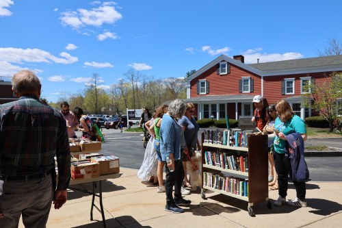 A group of people browsing books at an outdoor book sale on a sunny day, with a red building in the background.