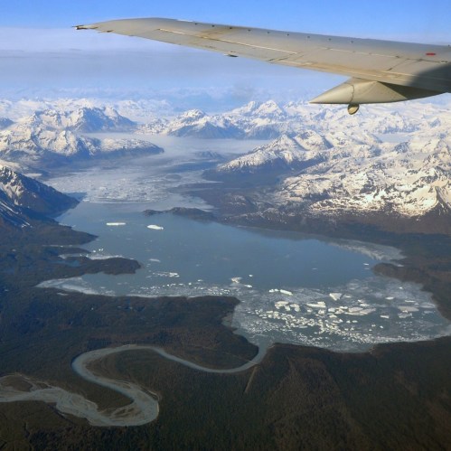 Aerial view of snow-capped mountains, a glacial lake, and winding rivers under a clear blue sky.