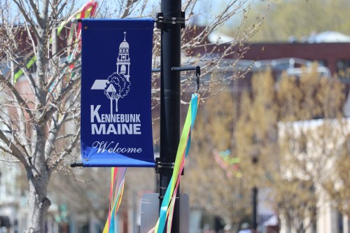 A blue banner reading "Kennebunk Maine Welcome" hangs from a pole, with colorful ribbons and trees in the background.