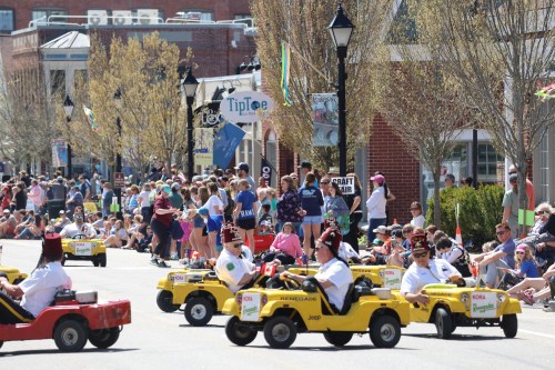 A lively parade scene with people watching as participants drive small yellow cars down the street.