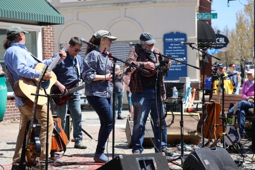 A lively outdoor music performance with musicians playing guitars and a banjo, surrounded by an audience enjoying the show.