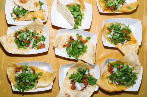 A variety of tacos on paper trays, filled with meat, onions, and fresh cilantro, arranged on a yellow table.