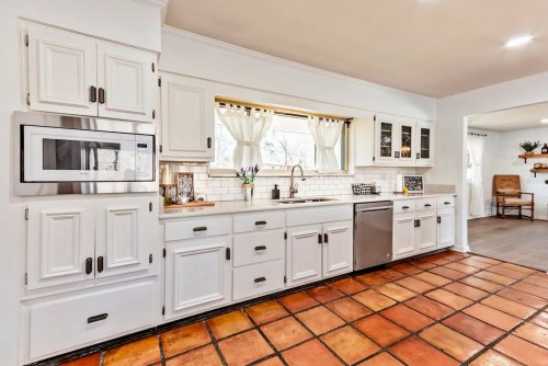 Bright kitchen with white cabinets, stainless steel appliances, and terracotta tile flooring. Natural light from a window.