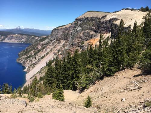 A scenic view of a rocky cliffside overlooking a blue lake, surrounded by green trees and distant mountains.