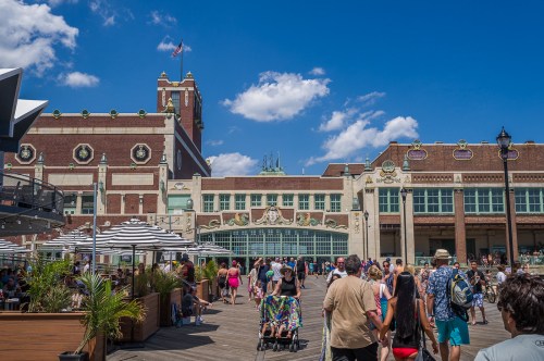 Crowd walking along a boardwalk with a historic building in the background under a bright blue sky.