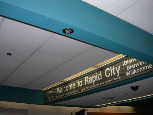 Sign welcoming visitors to Rapid City, featuring text in English and multiple languages, with a modern ceiling design.