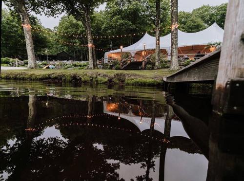 A serene outdoor scene featuring a tent, trees, and string lights reflected in calm water.