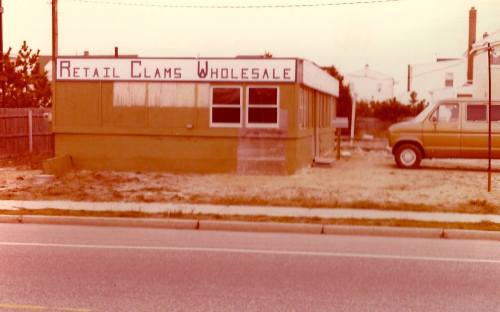 A vintage building with a sign reading "Retail Clams Wholesale," parked van, and gravel lot beside a road.