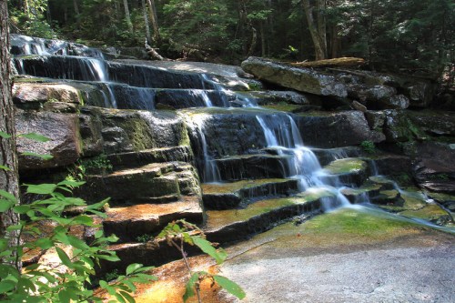 A serene waterfall cascades over rocky steps, surrounded by lush green trees and natural scenery.