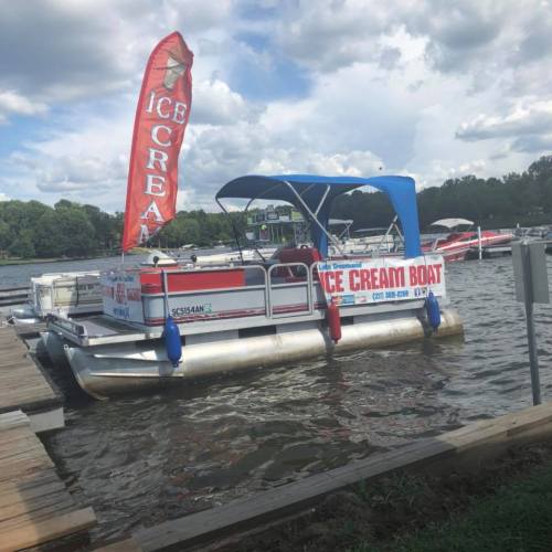 A pontoon boat with an "Ice Cream Boat" banner docked at a marina, surrounded by other boats and water.