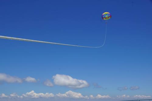 A colorful parachute floats high in a clear blue sky, tethered by a long rope against fluffy white clouds.
