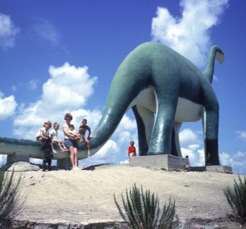 A group of children and adults pose around a large green dinosaur statue under a blue sky with fluffy clouds.