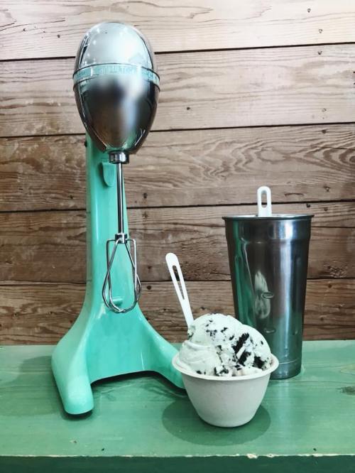 A vintage mint green mixer beside a bowl of cookies and cream ice cream and a metal cup on a wooden surface.