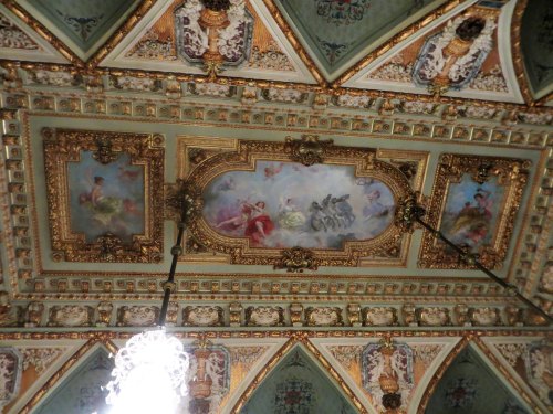 Ornate ceiling with intricate gold details and painted scenes, featuring a chandelier in the center.