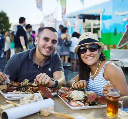 A smiling couple enjoys a seafood feast at an outdoor festival, with crabs and drinks on their table.