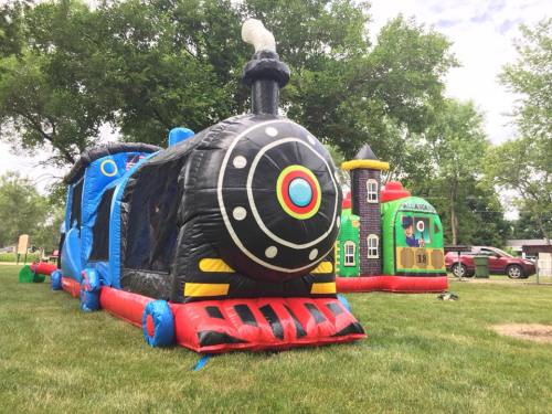 Two colorful inflatable bounce houses: a black train and a castle, set on green grass under a clear sky.